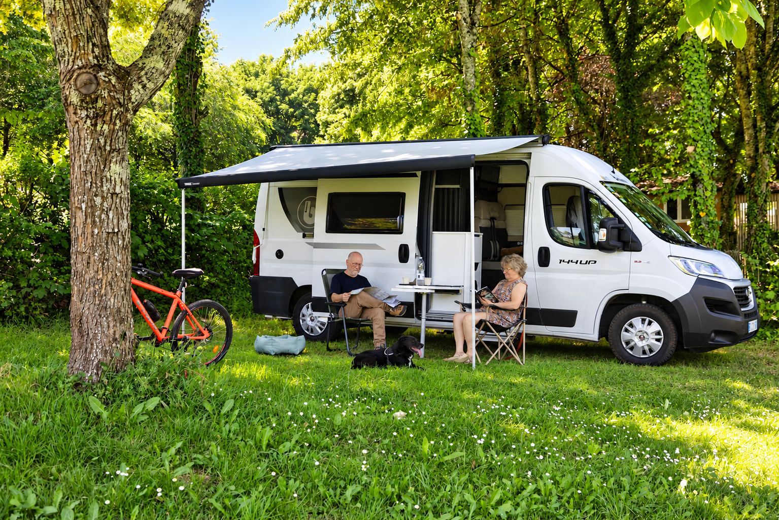 Un couple est assis sous l'auvent d'un van aménagé blanc, avec un chien noir et un vélo orange, dans une clairière herbeuse ombragée.