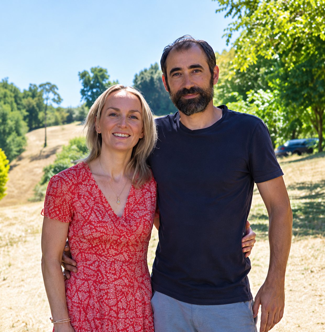 Une femme en robe rouge à motifs et un homme barbu en t-shirt sombre posent côte à côte, souriants, devant un paysage verdoyant ensoleillé.