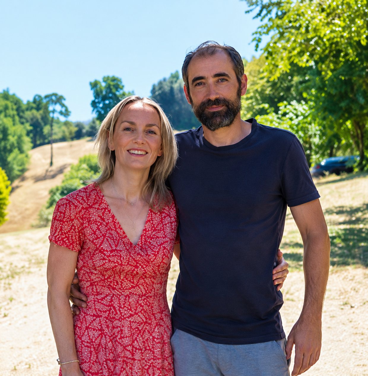 Une femme en robe rouge à motifs et un homme barbu en t-shirt noir posent côte à côte, souriants, devant des arbres et une colline en plein soleil.