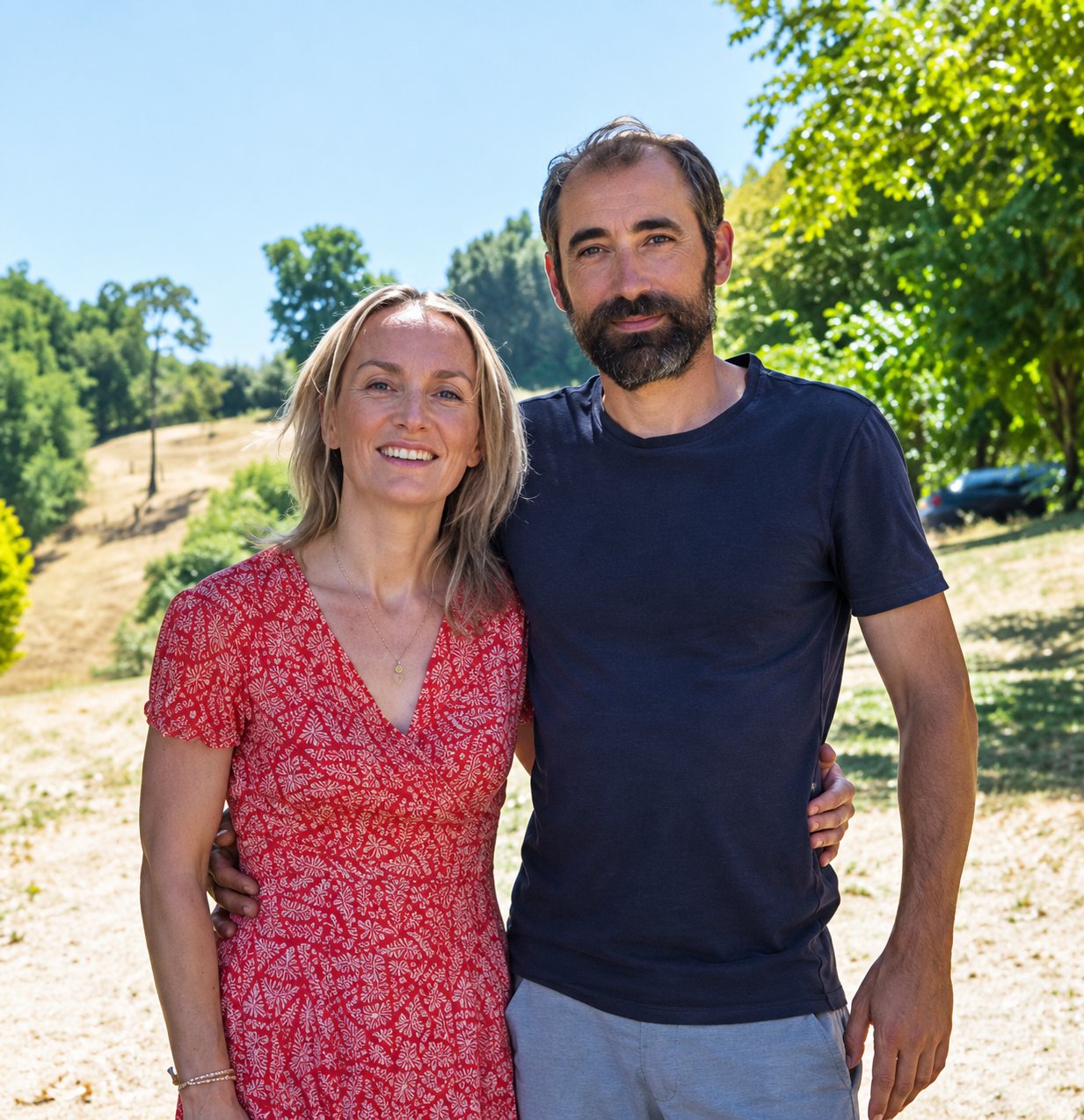 Une femme en robe rouge à motifs et un homme barbu en t-shirt noir posent côte à côte en plein air, devant des arbres et une colline ensoleillée.