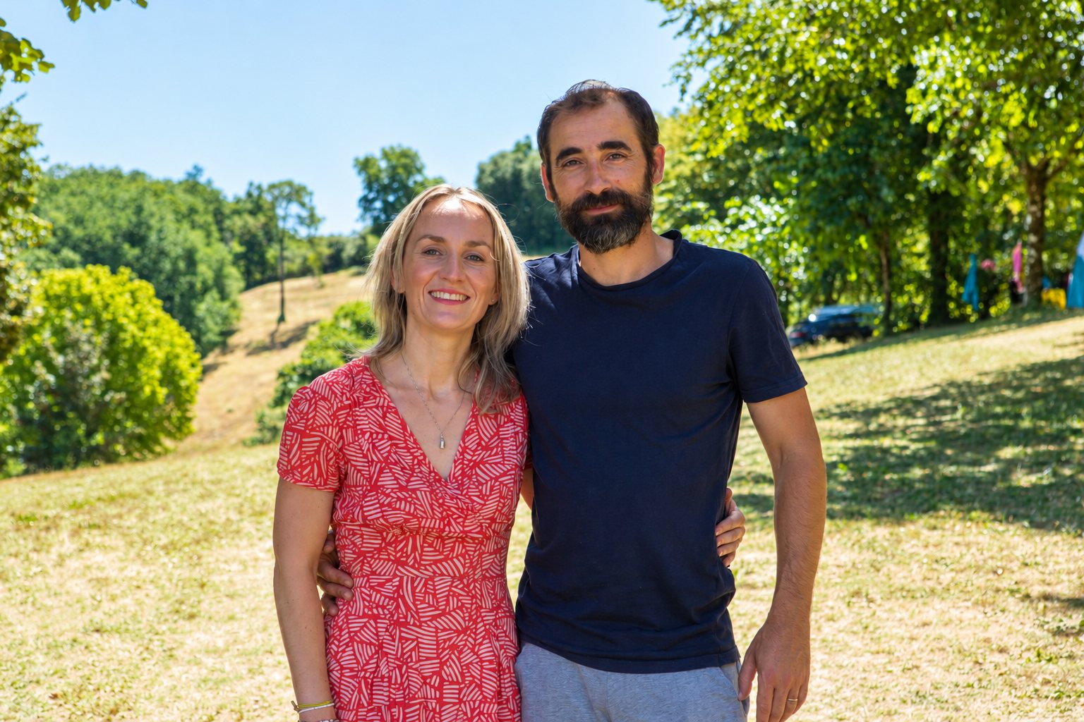 Une femme en robe rouge et un homme barbu en t-shirt sombre posent côte à côte, souriants, dans un parc ensoleillé entouré d'arbres.