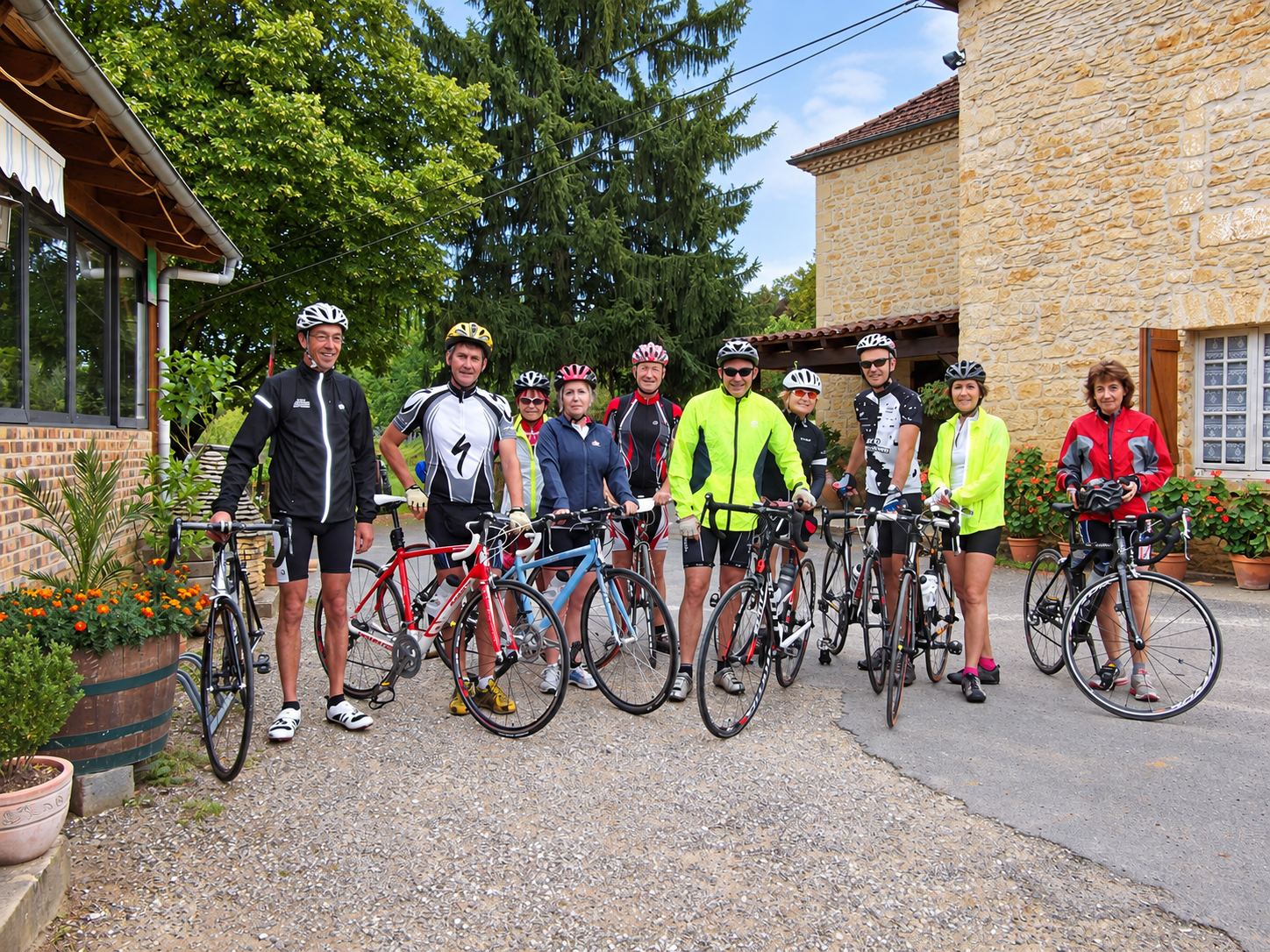 Un groupe de neuf cyclistes en tenue et casque pose avec leurs vélos de route sur un chemin devant une maison en pierre entourée de fleurs.