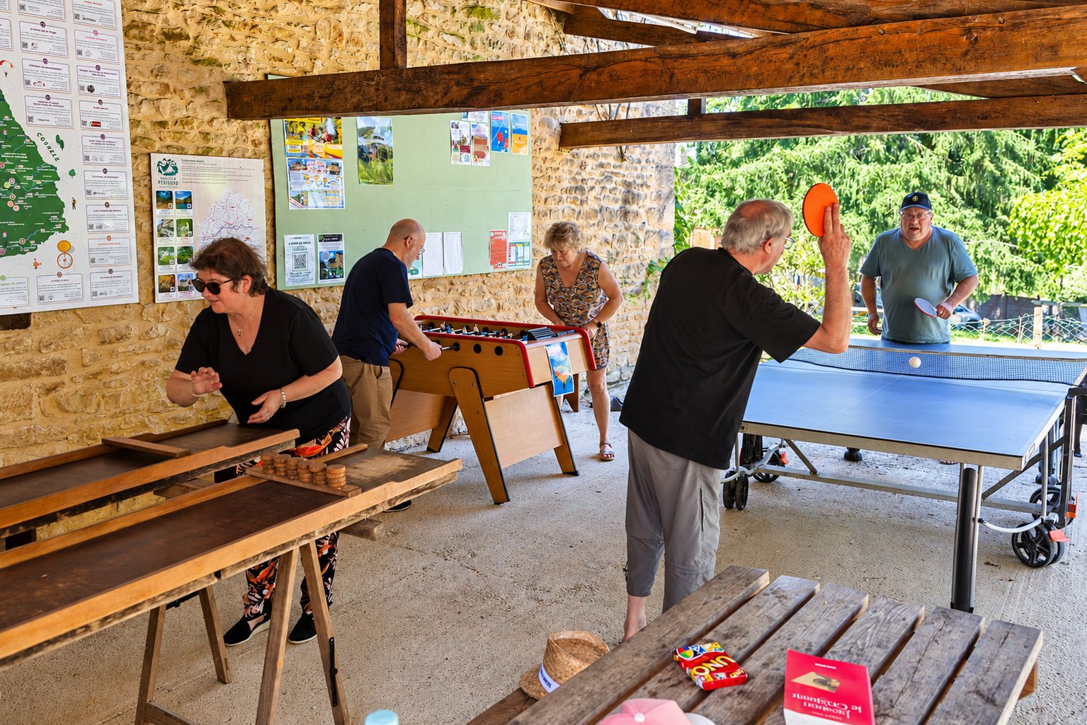 Plusieurs personnes jouent au ping-pong, au baby-foot et au jeu de palets sous un préau en pierre avec poutres en bois.