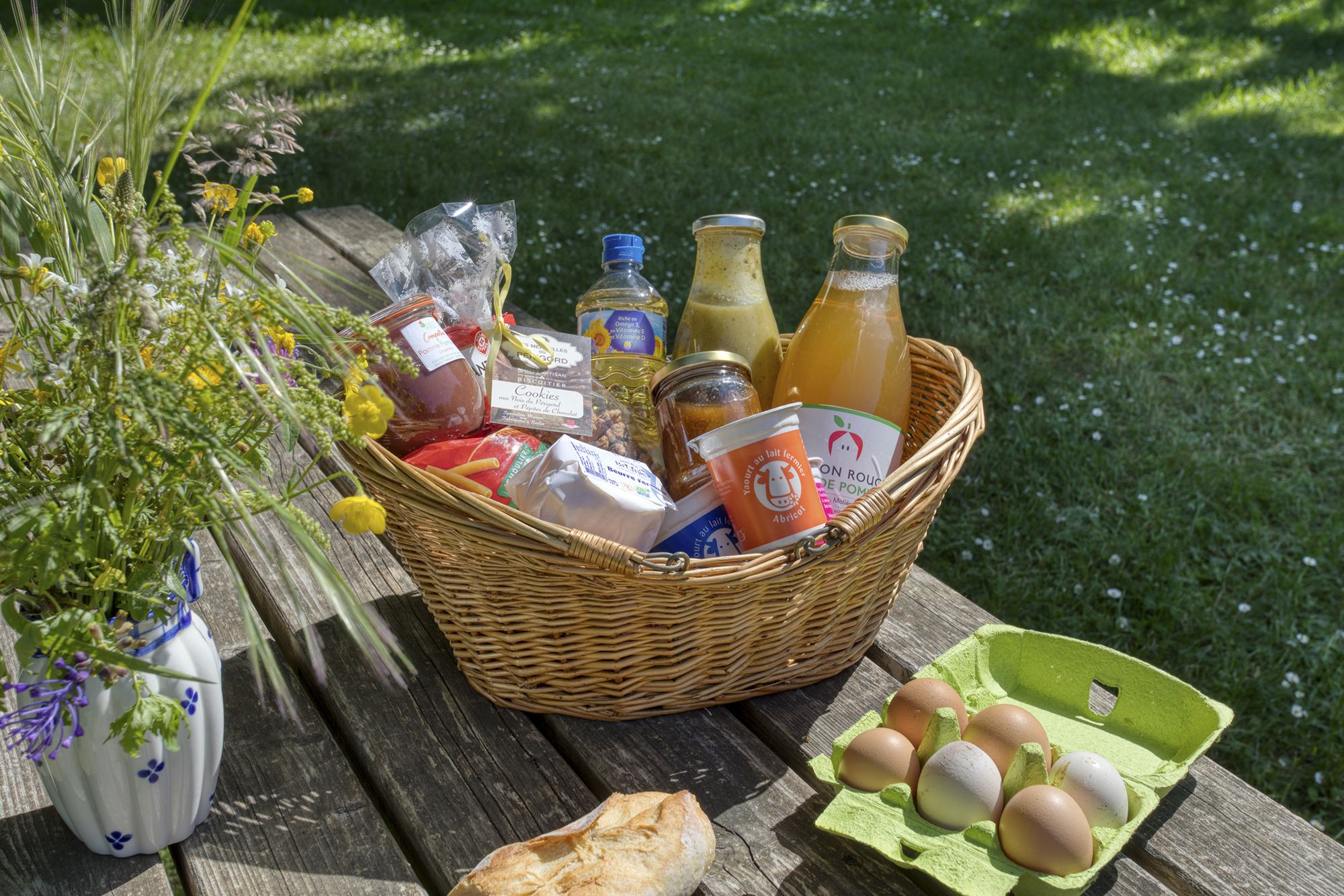 Un panier en osier rempli de produits du terroir, avec jus de fruits, yaourt, confiture et cookies, posé sur une table en bois près d'un bouquet de fleurs, d'œufs et d'un pain.